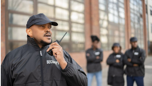 Agent de sécurité en uniforme noir parlant dans un talkie-walkie, avec une équipe de sécurité en arrière-plan, devant un bâtiment vitré